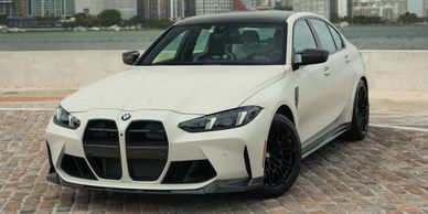 White BMW M3 with large black grille and black wheels parked on a brick surface.