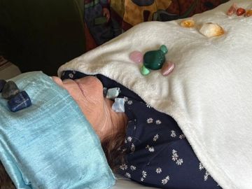 Person lying down with healing crystals on body and face, covered with a blanket.
