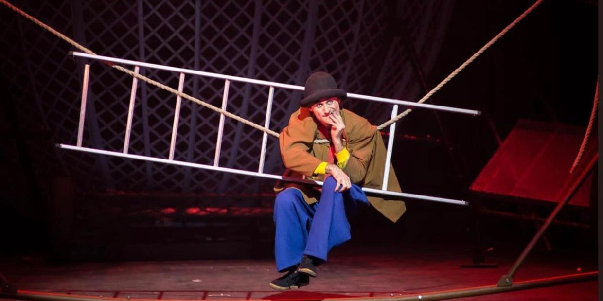 A clown in colorful clothes sits on a suspended ladder on stage, holding a finger to his lips.
