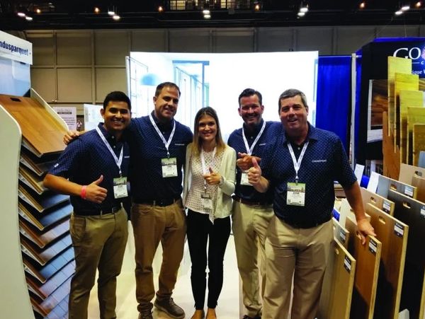 Five people at a trade show booth giving thumbs up, surrounded by wooden flooring samples.