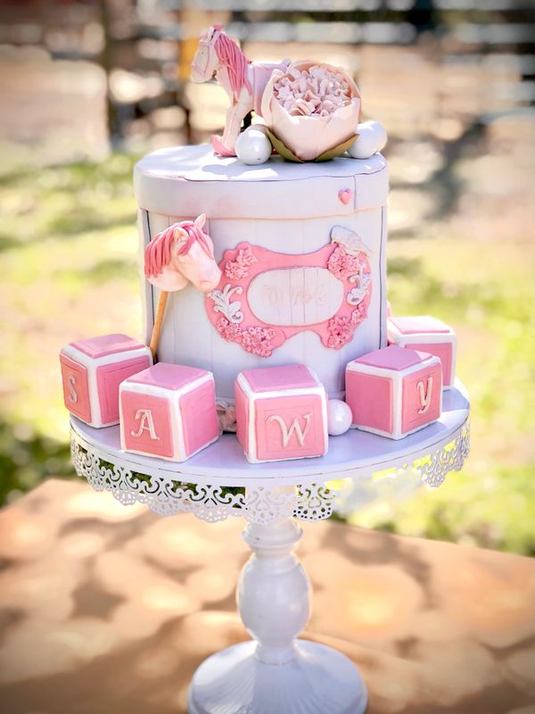 Elegant pink and white baby shower cake with horse and floral decorations.