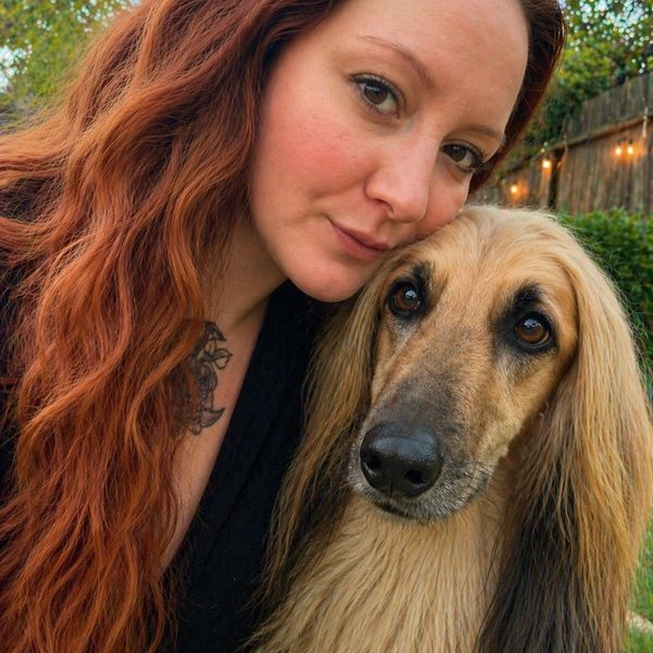 Woman with long red hair posing closely with a long-haired dog outdoors.