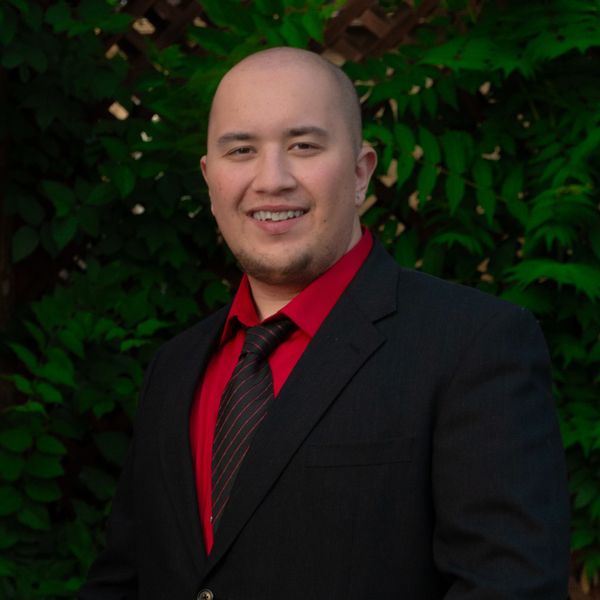 Man in a black suit and red shirt smiling outdoors.
