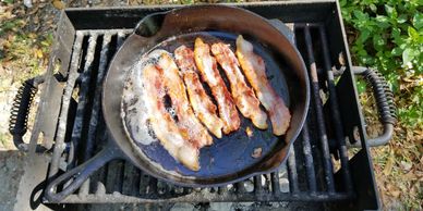 6 pieces of bacon sizzling in the sunlight in a cast iron skillet over a campsite grill. 