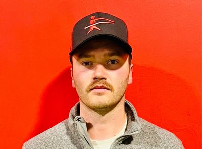 Man with mustache wearing a black cap and gray jacket against a red background.
