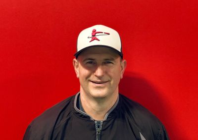 Smiling man in a black jacket and white cap stands against a red background.