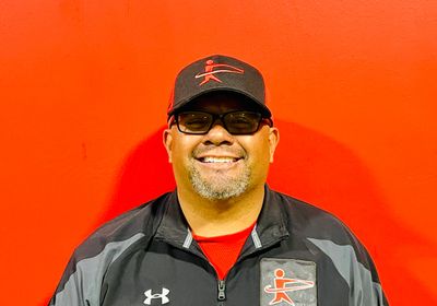 Smiling man in sporty black jacket and cap against a bright red background.