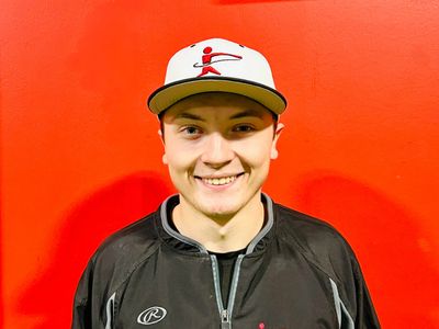 Young man smiling in a black jacket and white cap against a red wall.