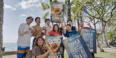 Group of friends holding promotional Shaka posters outdoors near the beach.