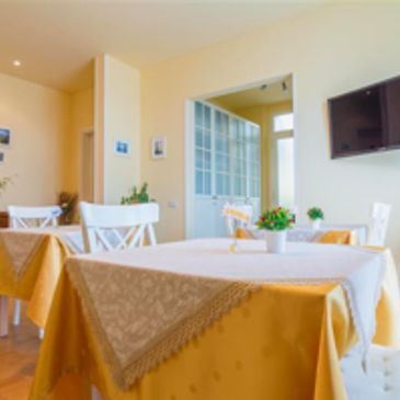 Bright dining room with yellow tablecloths and white chairs, featuring a wall-mounted TV.
