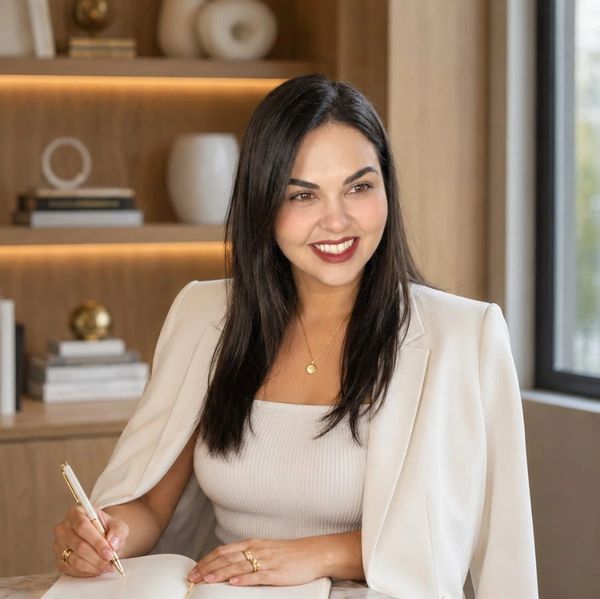 Smiling woman in white blazer writing in a notebook at a wooden desk.