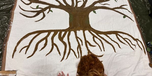 Person with red hair painting a large tree mural on a white canvas.