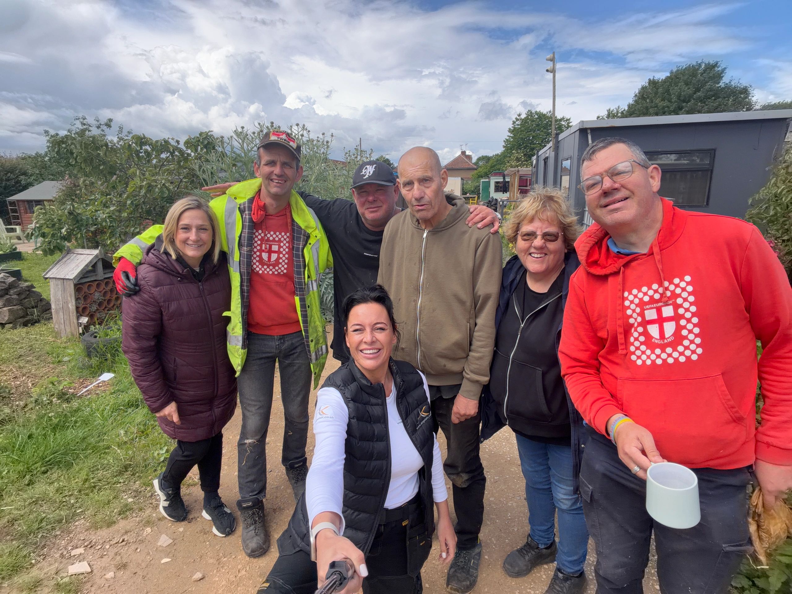 A cheerful group selfie of seven people outdoors on a cloudy day.