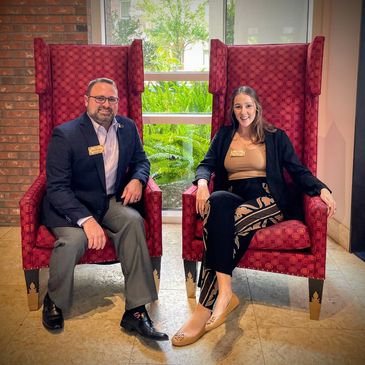 Two professionals sitting on red armchairs by a window with greenery outside.