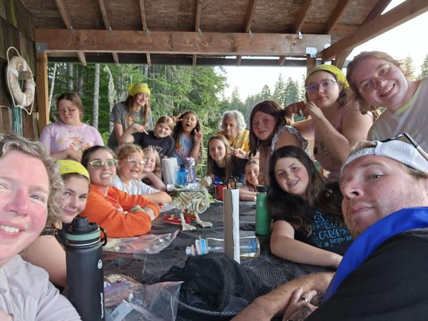 A large group of people gathered around a table outdoors under a wooden roof.