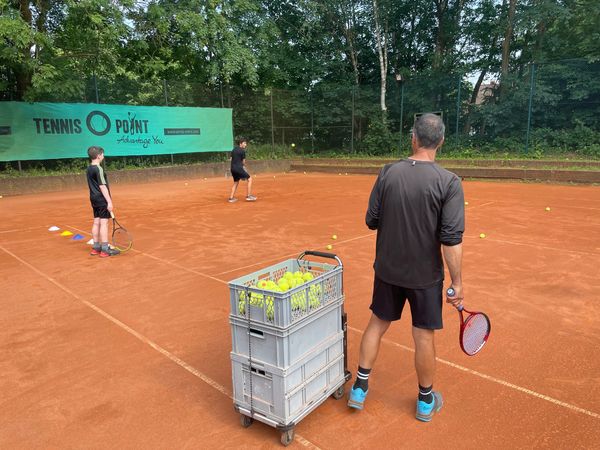 A tennis coach instructs two young players on a clay court with many tennis balls scattered.