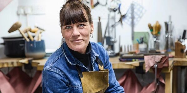 Woman in a workshop wearing a denim jacket and apron, smiling at the camera.