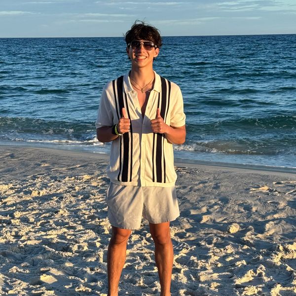 A young man smiling on the beach with thumbs up, wearing sunglasses and casual summer clothes.
