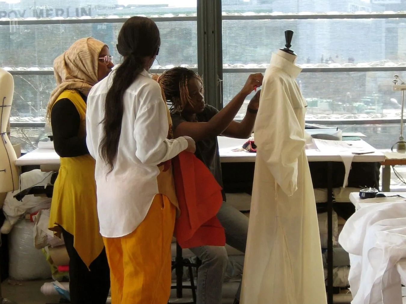 Three women work together on designing a garment on a mannequin in a bright studio.
