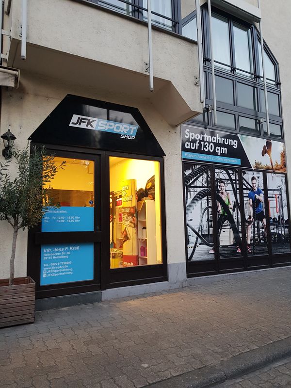Eingang zum JFK Sport Shop mit Sportnahrung-Werbung.