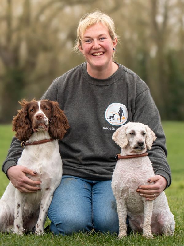 Smiling woman kneels on grass with two dogs, one brown-and-white and one curly white.