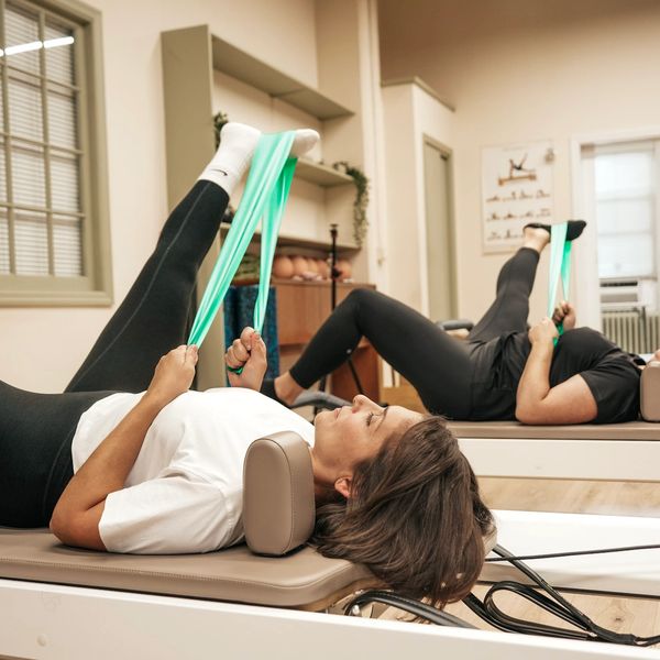 Two people doing resistance band leg stretches in a pilates studio.