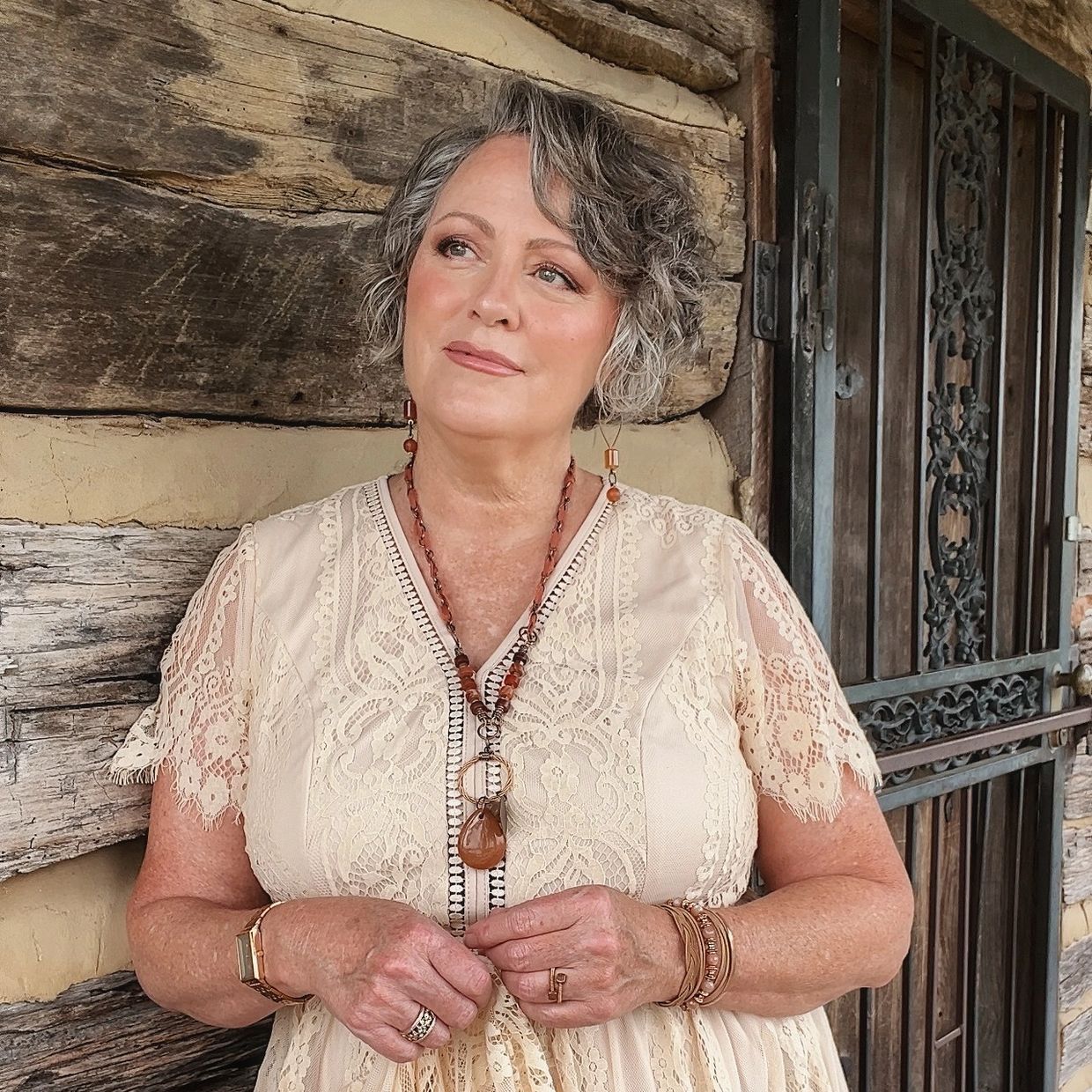 Woman in a cream lace dress with jewelry, standing by a rustic wooden wall.