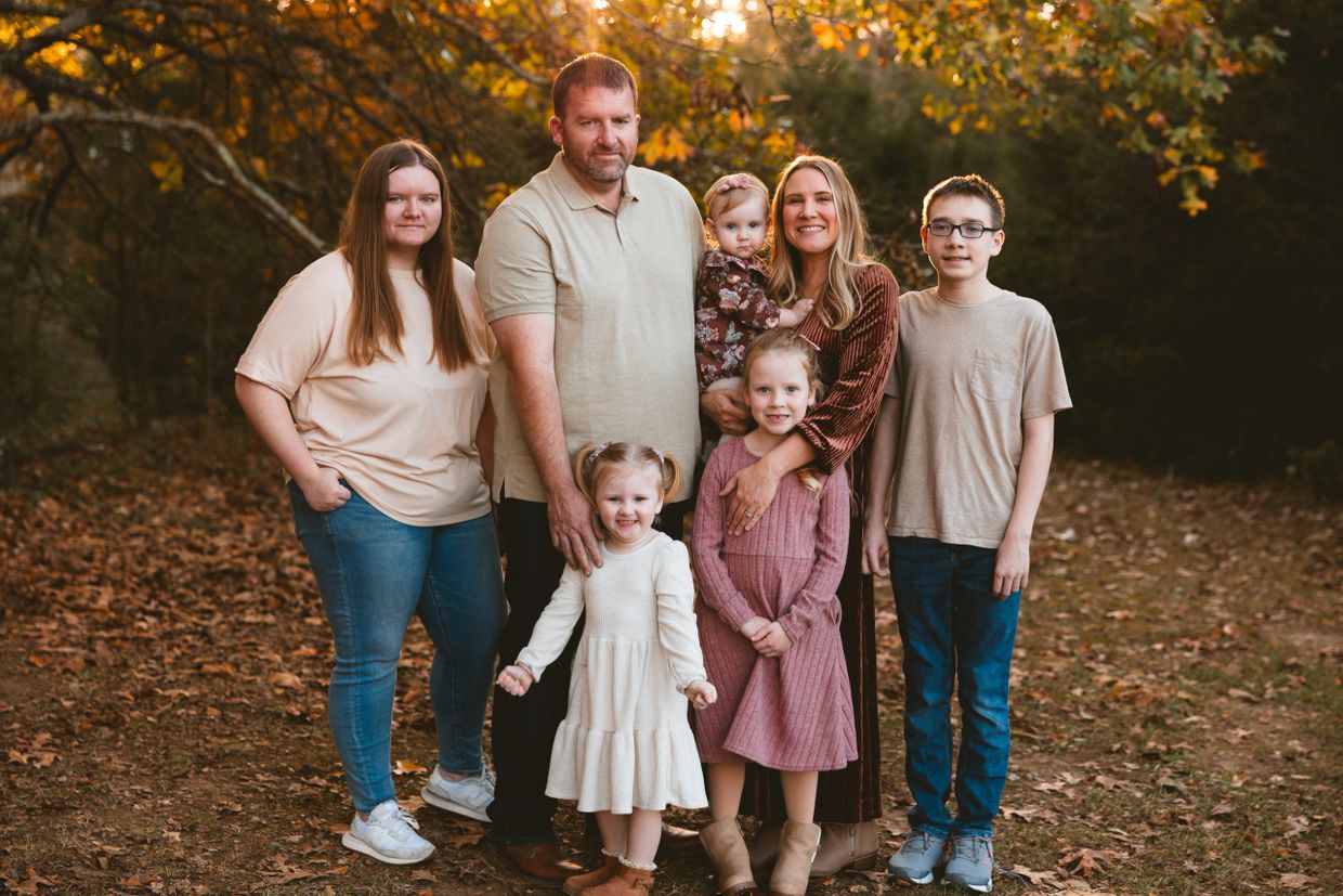 A family of seven posing outdoors during autumn with warm sunlight and colorful leaves.
