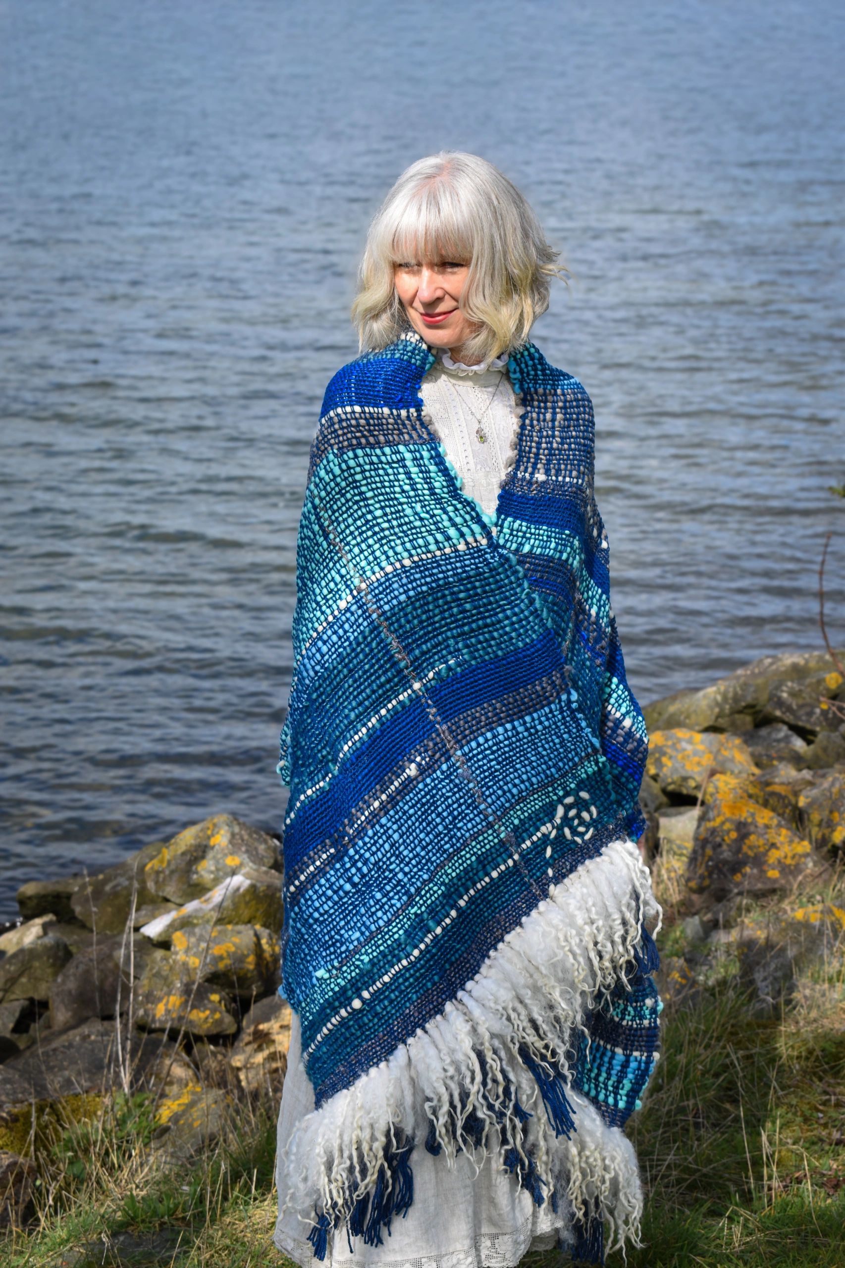 Woman wrapped in a blue and white shawl by the water, standing on rocky ground.