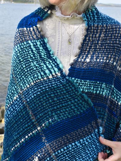 Person wrapped in a blue and gray woven shawl by the water.