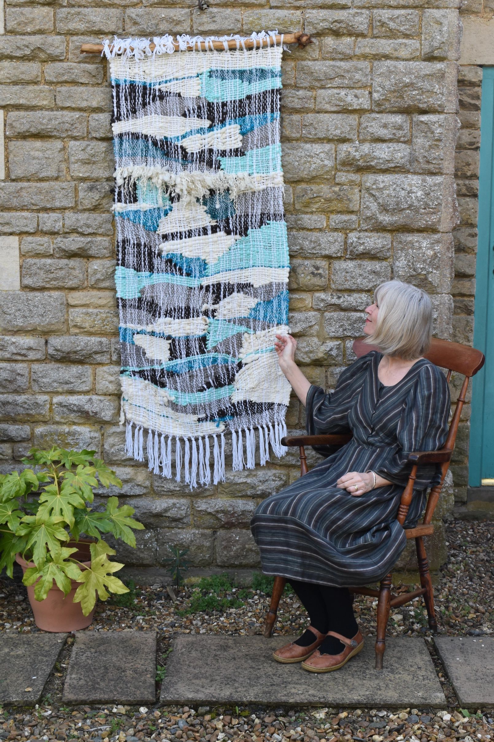 Woman sitting on a wooden chair beside a woven wall hanging on a stone wall.