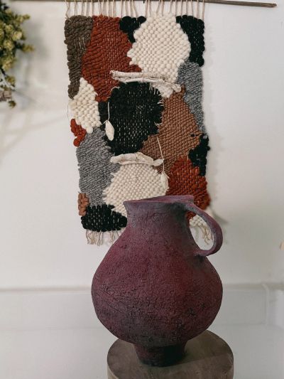 Rustic textured vase in front of a colorful woven wall hanging.