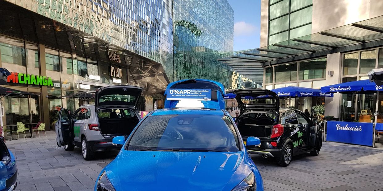 Blue car with open trunk in urban plaza near restaurants and shops.