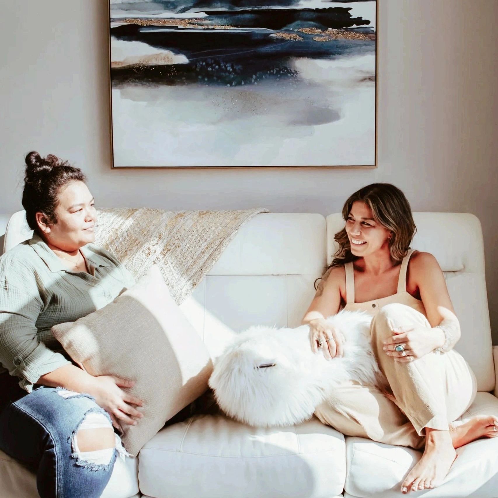 Two women sitting and chatting on a white couch in a cozy living room.
