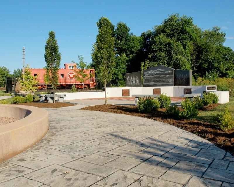 Memorial park with a red train caboose and monument surrounded by trees and a paved walkway.