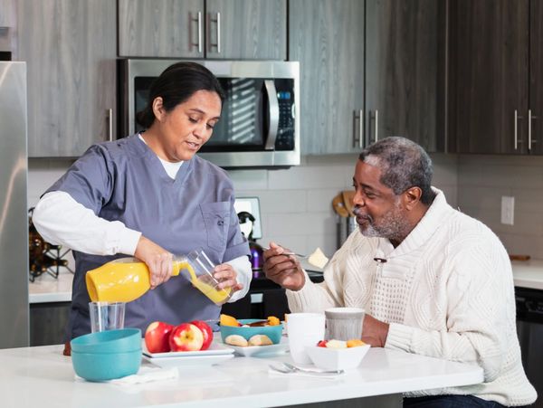 A healthcare professional in gray scrubs pours orange juice into a glass for an elderly man.