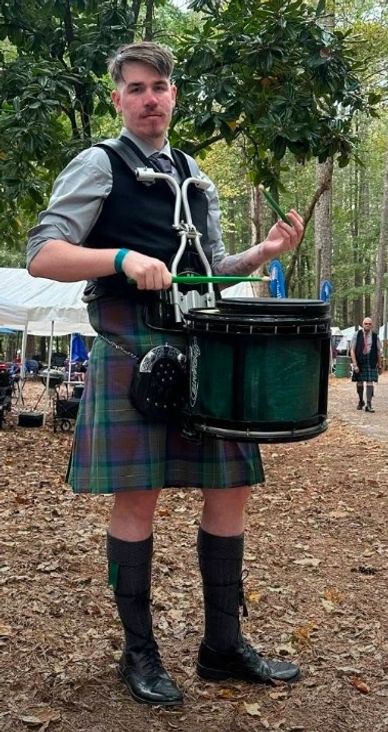 Man in a kilt playing a snare drum outdoors.