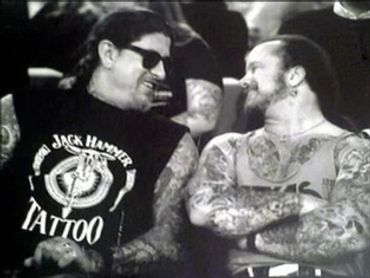 Two tattooed men smiling and talking, one wearing sunglasses.