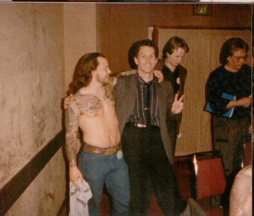 Four men posing casually in a dimly lit room with one shirtless and tattooed.