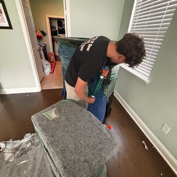A man wrapping furniture in protective blankets with plastic wrap indoors.