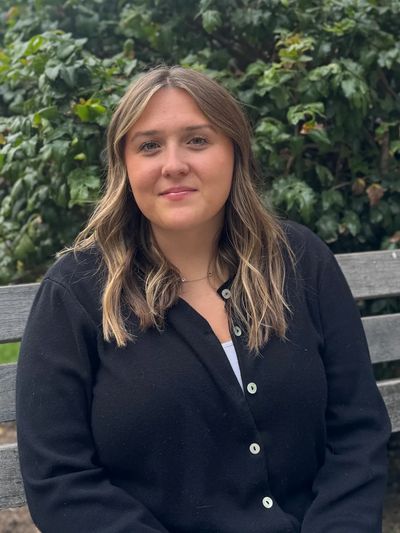 A woman sitting on a bench outdoors in front of greenery.