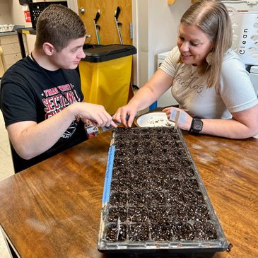 Two people planting seeds in a seed tray indoors, smiling and engaged.