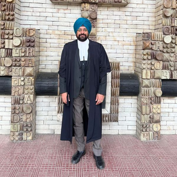 Man in traditional lawyer attire and blue turban standing before a decorative stone wall.