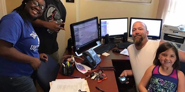 Four people smiling in an office with multiple computer screens and a U.S. map on the wall.