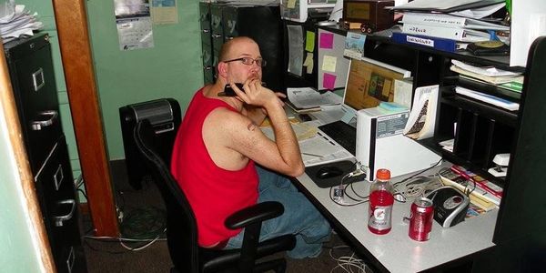 Man in red tank top working at cluttered office desk with computer and papers.