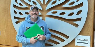 Man in blue hoodie holding green folder, smiling near elevator sign and tree wall art.