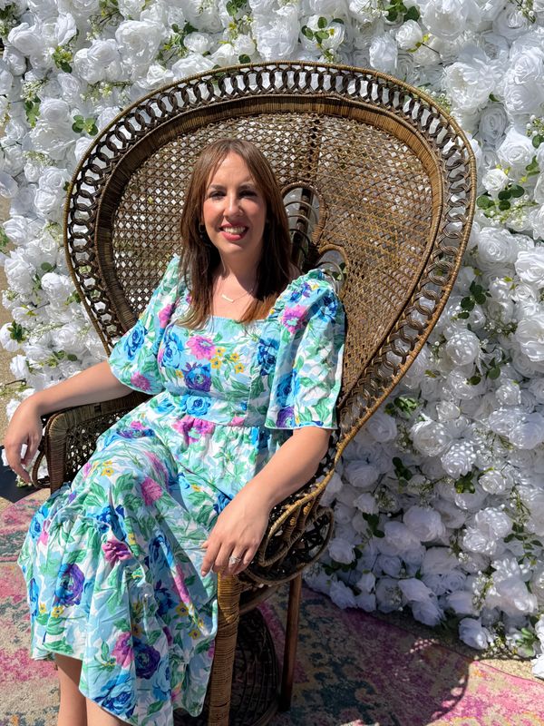 Woman in floral dress sitting on wicker chair against white flower wall.