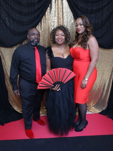 Three elegantly dressed people posing on a red and black carpet with a glittery backdrop.