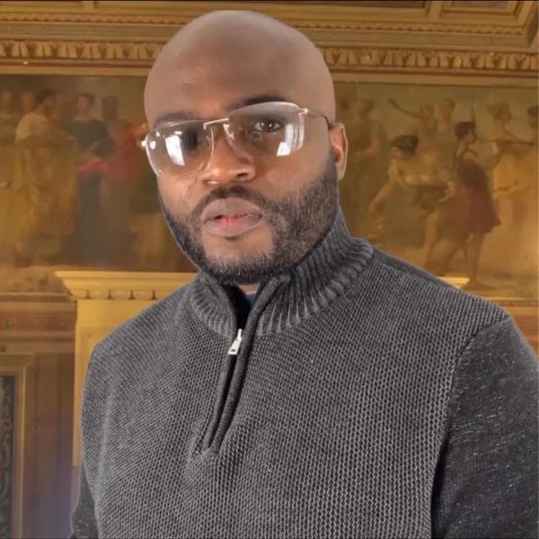 Man wearing glasses and a gray sweater in a room with classical paintings.