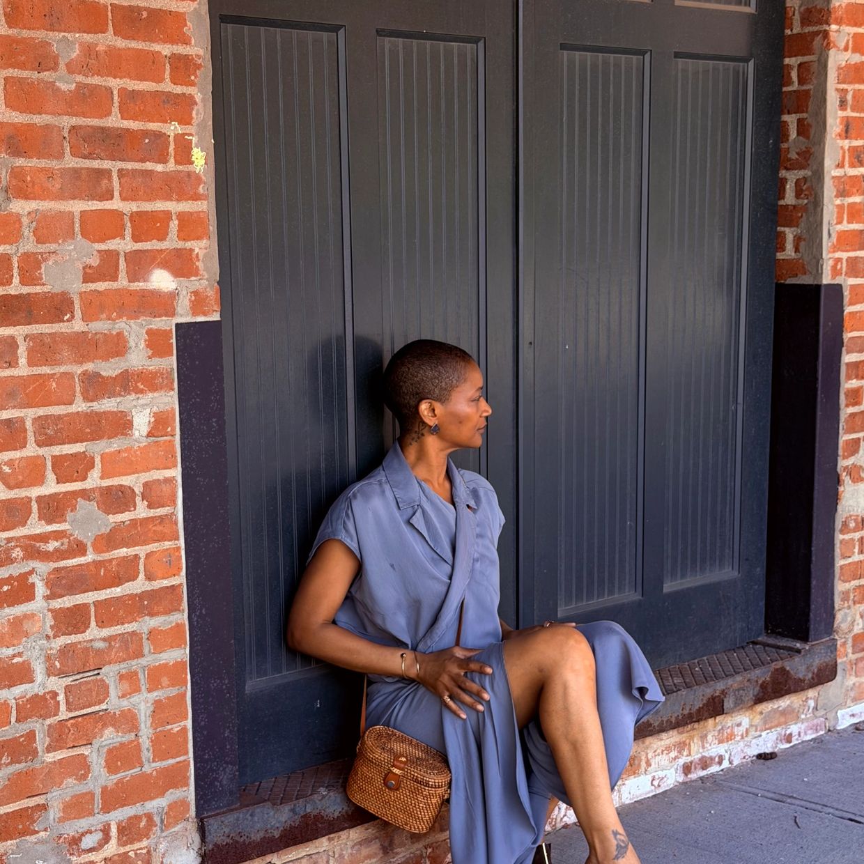 Woman in gray dress sitting against black panels and brick wall.